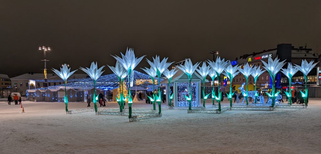 In this photo is shown the snowy Kuopio market square adorned with tall flowerlike lighting fixtures. In the background can slightly be seen the blue lights covering the Christmas market.